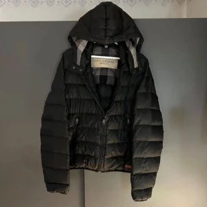 Svart dunjacka från Burberry - Snygg svart dunjacka från Burberry med huva och dragkedja. Jackan har ett quiltat mönster och två fickor med dragkedjor. Perfekt för kyliga dagar. Perfekt för värdet nu och framöver. Storlek M