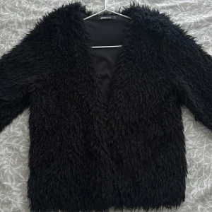 Svart pälsjacka från Gina Tricot - Snygg svart pälsjacka från Gina Tricot. Jackan har en fluffig textur och är perfekt för att hålla dig varm och stilren. Den har långa ärmar och en öppen design utan knappar eller dragkedja. Knappt använd