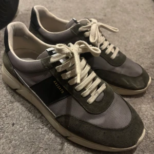 Grå sneakers från Axel Arigato - Snygga grå sneakers från Axel Arigato med vita snören och svarta detaljer. Tillverkade i Portugal, dessa skor kombinerar stil och komfort perfekt.