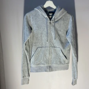 Grå hoodie från Juicy Couture - Säljer en stilren grå hoodie från Juicy Couture med dragkedja och huva. Tröjan har en broderad logga på bröstet och praktiska fickor framtill. Perfekt för en avslappnad look. Förlåt för suddig bild