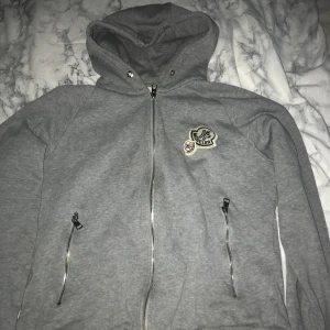 Grå hoodie från Moncler - Säljer en stilren grå hoodie från Moncler med dragkedja och två fickor framtill. Den har en broderad logga på bröstet och en bekväm huva. Perfekt för en avslappnad look.