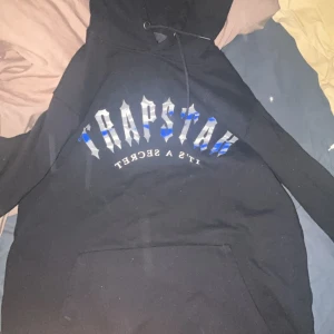 Svart hoodie från Trapstar - Säljer en svart hoodie från Trapstar med tryck på både fram- och baksidan. Framsidan har texten 'Trapstar It's a Secret' i vitt och blått, medan baksidan har ett grafiskt tryck med texten 'Global Ties'. Hoodien har en klassisk känguruficka och justerbar huva.