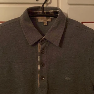 Grå pikétröja från Burberry - Säljer en stilren grå pikétröja från Burberry med klassisk krage och knappar framtill. Tröjan har långa ärmar och ett diskret broderat emblem på bröstet. Perfekt för en elegant och avslappnad look.