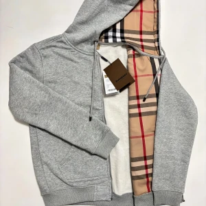 Grå hoodie från Burberry - Säljer en stilren grå hoodie från Burberry med klassiskt rutigt foder i huvan. Tröjan har en broderad detalj på bröstet och dragkedja framtill. Storleken är M och kan även passa någon med S. Skickas inom 24 timmar.