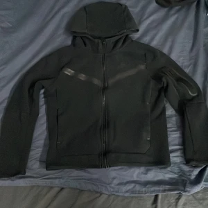 Svart huvtröja från Nike - Säljer en svart huvtröja från Nike med dragkedja och huva. Tröjan har en standard passform och är tillverkad i ett syntetmaterial. Perfekt för en sportig look.