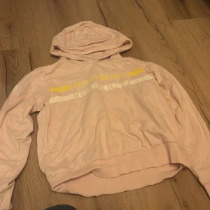 Rosa hoodie från Hampton Republic - Säljer en rosa hoodie från Hampton Republic med tryck i vitt och gult på bröstet. Tröjan har en bekväm huva och långa ärmar, perfekt för en avslappnad stil.