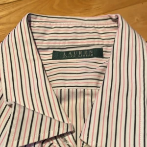Randig skjorta från Lauren Ralph Lauren - Snygg randig skjorta från Lauren Ralph Lauren i vitt med tunna ränder i rosa och mörkblått/svart. Skjortan har långa ärmar och knappar framtill. Perfekt för en stilren look. Hittar ingen storlek men gissar på 44.