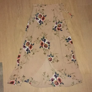 Beige blommig omlottkjol - Charmig beige omlottkjol med blommigt mönster i rosa och blått. Kjolen har en knytning i midjan och en elegant slits framtill. Perfekt för en somrig look! Dock en liten defekt vid knytningen, men går enkelt att sy!