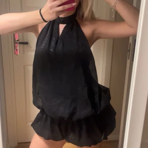 Svart ärmlös klänning - Snygg svart ärmlös klänning med halterneck och volangdetalj. Den har en öppen rygg och en luftig passform, perfekt för en kväll ute.
