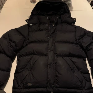 Svart/marinblå dunjacka från Morris - Säljer en svart/marinblå dunjacka från Morris med huva och dragkedja. Jackan har dragkedja och knappar framtill. Perfekt för kalla dagar, då det är dun och fjäder fyllning.