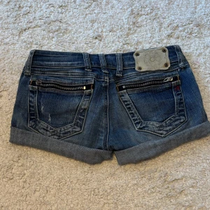 Lågmidjade jeansshorts - Snygga blå jeansshorts från Replay med coola dragkedjedetaljer på bakfickorna. De var tidigare jeans men den tidigare ägaren klippte till shorts. Passar som minishorts med en låg midja, kontakta för bilder på. Midjan: 36cm höft: 41cm längd: 24cm