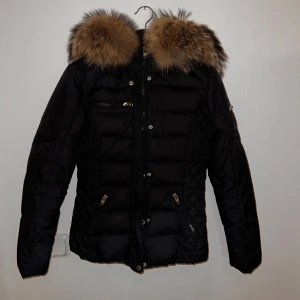 Svart dunjacka med päls - Snygg svart dunjacka från Rock and Blue med en fluffig pälsdetalj på huvan. Jackan har dragkedjor och knappar framtill samt flera praktiska fickor. Perfekt för kalla vinterdagar. Äkta päls på luvan❤️tar emot bud. Två defekter -SE bild