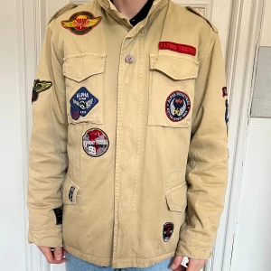 Alpha Industries M65 Field Jacket - Säljer en beige jacka från Alpha Industries med flera coola patches. Jackan har långa ärmar och knappar framtill. Perfekt för vår och höst. Storlek L. Se mina övriga annonser för fler grymma plagg! 