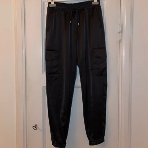 Svarta satin cargopants - Snygga svarta cargopants med elastisk midja och praktiska fickor på sidorna. Perfekta för en avslappnad stil med en touch av funktionalitet. Passar bra till sneakers eller kängor.