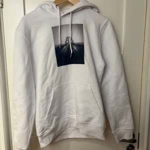 Vit hoodie med tryckt motiv - Säljer en stilren vit hoodie med ett coolt broderat motiv av en bro på framsidan. Hoodien har en klassisk passform med känguruficka och justerbar huva med dragsko. Perfekt för en avslappnad look.