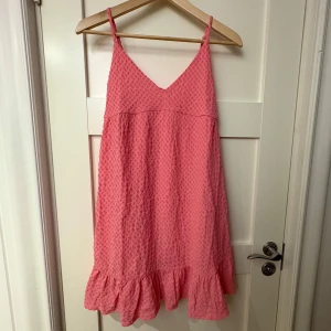 Rosa klänning från Gina Tricot - Säljer en söt rosa klänning från Gina Tricot med smala axelband och volangdetalj vid fållen. Klänningen har en v-ringad design och ett texturerat mönster som ger en fin touch. Perfekt för varma dagar!