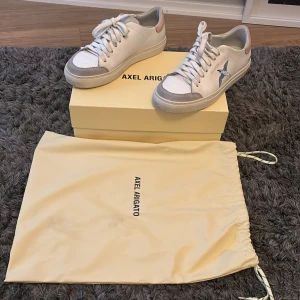 Vita sneakers från Axel Arigato - Snygga vita sneakers från Axel Arigato med en blå fågelbrodering på sidan. Skorna har snörning och en beige detalj vid hälen. Perfekta för en stilren look. Jättesköna, säljer för att jag nu inte längre kan ha dem. Tror även att dem slutat säljas.
