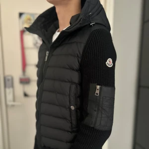 Svart cardigan från Moncler - Snygg svart cardigan från Moncler med stickade ärmar och dragkedja framtill. Cardigan har en broderad Moncler-logga på ärmen och en praktisk ficka med dragkedja. Perfekt för kyligare dagar.