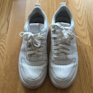 Vita sneakers från Hugo - Snygga vita sneakers från Hugo med klassisk snörning och stilren design. Skorna har en tydlig logga baktill och är tillverkade i syntetmaterial. Perfekta för en casual look.