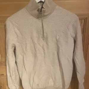 Polo Tröja - Säljer en stilren beige stickad tröja med halv dragkedja och hög krage. Perfekt för kyligare dagar. Tröjan har långa ärmar och en diskret logga på bröstet. Passar både till vardags och mer uppklädda tillfällen.