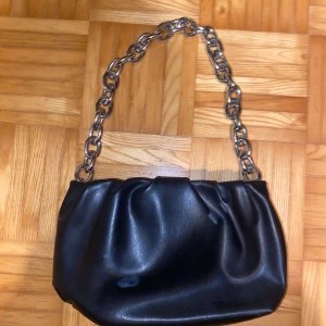 Svart axelväska med kedjehandtag - Snygg svart axelväska i skinn med ett coolt kedjehandtag i metall. Väskan har en dragkedja upptill för säker förvaring. Ställ gärna frågor!😊