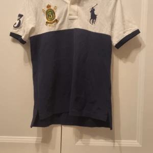 Vit och blå pikétröja med broderad logga - Säljer en stilren vit och blå pikétröja från Ralph Lauren. Tröjan har korta ärmar och en broderad logga på bröstet. Perfekt för en avslappnad men ändå snygg look. Storlek S.