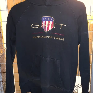 Svart hoodie från GANT - Säljer en svart hoodie från GANT med broderad logga och texten 'American Sportswear' på framsidan. Tröjan har en klassisk känguruficka och en bekväm huva. Perfekt för en avslappnad stil. Barnstorlek M/ Motsvarande S i vuxen 