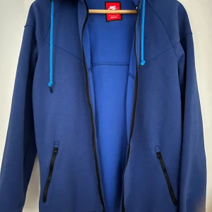 Blå hoodie från Nike - Säljer en snygg blå hoodie från Nike med dragkedja och huva. Den har långa ärmar och ribbade muddar. Perfekt för en casual look. Den har även två praktiska fickor med dragkedja framtill. Använd sparsamt, mycket gott skick! Storlek M. 