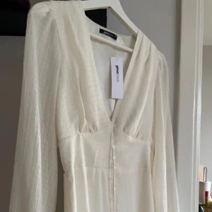 Vit klänning från Gina Tricot - Elegant vit klänning från Gina Tricot med långa ärmar och knappar framtill. Klänningen har en v-ringad design och är perfekt för somriga tillfällen. Den är tillverkad i ett lätt och luftigt material som ger en skön känsla.