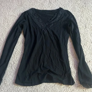 Svart långärmad topp med spetsdetaljer - Säljer en elegant svart långärmad topp med v-ringning och spetsdetaljer vid halsen och ärmsluten. Perfekt för en stilren look.