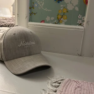 Grå keps från Norrland - Snygg grå keps från Norrland med broderad text och en liten orange detalj på sidan. Perfekt för en avslappnad stil. Justerbar passform baktill.