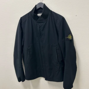 Svart jacka från Stone Island - Stone Island (RARE) Soft Shell-R Primaloft Insulation Technology  Ett högpresterande plagg känt för sina vattenavvisande, vindtäta och andningsbara egenskaper. 	•	Storlek: Herr L (Jag har använt den och är storlek XS / 163 cm – den sitter perfekt oversized på mig). 	•	Funktioner: Stor innerficka med plats för pass, telefon etc. med knappförslutning. 	•	Nypris: Ca 8 700 SEK. 	•	Pris: 2 100 SEK. 	•	Mötesplats: Kan mötas i innerstan eller skicka med post.