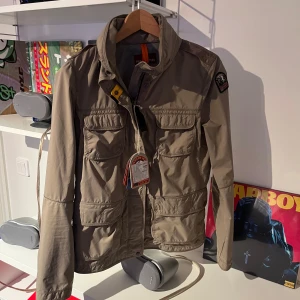 Jacka från Parajumpers - Säljer en beige jacka från Parajumpers med flera praktiska fickor och en dragkedja framtill. Jackan har en cool patch på ärmen och är perfekt för blåsiga dagar. Den har en stilren design med detaljer som knappar och en ribbad krage.