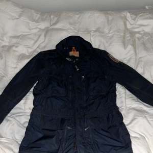 Mörkblå jacka från Parajumpers - Säljer en mörkblå jacka från Parajumpers med huva och flera praktiska fickor. Jackan har en dragkedja och knappar framtill samt en broderad logga på ärmen. Perfekt för kyligare dagar med sin stilrena design och funktionella detaljer. Kan gå ner i pris vid snabb affär.