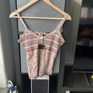 00s topp från Kookai - Säljer en unik topp från Kookai med färgglada ränder och blommigt mönster. Toppen har smala axelband och knappar framtill. Baksidan är ribbad i svart. Insydd av mig vid ryggen men går att klippa bort
