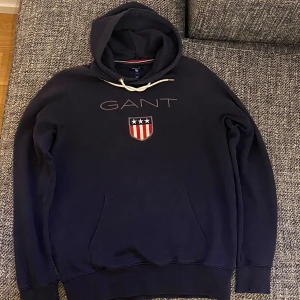 Mörkblå hoodie från GANT - Säljer en snygg mörkblå hoodie från GANT med logotyp och emblem på bröstet. Tröjan har en klassisk känguruficka och justerbar huva med snören. Perfekt för en avslappnad stil. 
