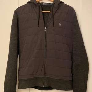 Svart cardigan från Polo Ralph Lauren - Säljer en svart jacka från Polo Ralph Lauren med dragkedja och huva. Jackan har en quiltad framsida och långa ärmar i ett mjukt material. Perfekt för kyligare dagar.