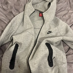 Grå hoodi Niketec - Säljer en grå hoodie från Nike med dragkedja och huva. Jackan har svarta detaljer och två fickor med dragkedjor. Perfekt för en sportig look eller en avslappnad dag. Det är en liten fläck på den 