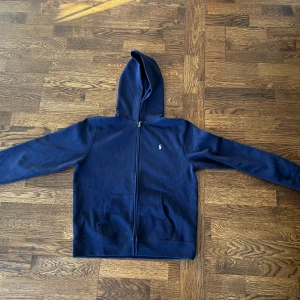 Mörkblå hoodie från Ralph Lauren - Säljer en snygg mörkblå hoodie från Ralph Lauren med dragkedja och två fickor framtill. Perfekt för en avslappnad stil. Den har en liten broderad logga på bröstet och är i storlek XS. Passar som en S dock. Helt oanvänd, har däremot inte kvar prislappen. Den är därför inte markerad som ny.