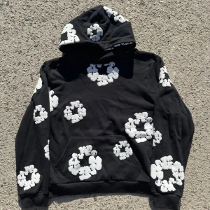 Denim Tears Hoodie - Säljer en cool svart hoodie från Denim Tears med vita blommor över hela tröjan. Den har en klassisk känguruficka och en bekväm huva. Perfekt för att sticka ut i mängden!