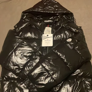 Svart dunjacka från Moncler - Säljer en snygg svart dunjacka från Moncler. Jackan har en glansig finish och är perfekt för kalla dagar. Den har en dragkedja framtill och en klassisk Moncler-logga på bröstet. Perfekt för att hålla dig varm och stilren under höst/vinter. Helt ny o välbehållen inte riktigt min stil bara.