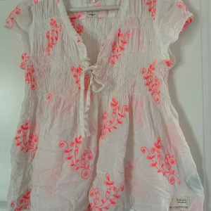 Vit blus med broderi från Odd Molly - Säljer en söt vit blus från Odd Molly med neonrosa broderade detaljer. Blusen har korta ärmar och en knytning framtill. Perfekt för en bohemisk look med sina fina broderier och luftiga passform. Toppen är i fint skick och passar perfekt till vår/sommar garderoben! Vid frågor eller funderingar hör av er, jag svarar så fort jag kan!🩷