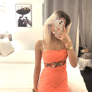 Orange klänning med mönstrad midja - Snygg orange klänning med en unik mönstrad detalj i midjan. Klänningen har en tight passform och är ärmlös med smala axelband. Perfekt för att sticka ut med sin livfulla färg och stilrena design.