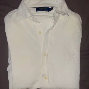 Vit stickad piké tröja från Ralph Lauren - Säljer en klassisk vit stickad piké tröja från Ralph Lauren med broderad logga på bröstet. Skjortan har långa ärmar och knappar framtill. Perfekt för en stilren look.