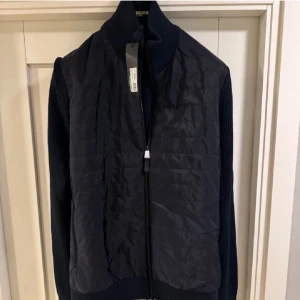 Mörkblå j.lindeberg cardigan - Snygg mörkblå cardigan från j.lindeberg med dragkedja framtill. Västen har en stilren design och är perfekt för lager-på-lager. Den har ribbstickade detaljer på sidorna och en hög krage för extra värme.