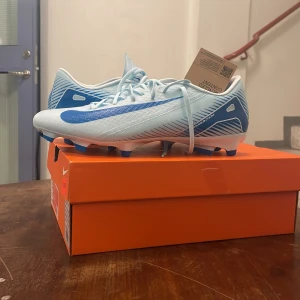 Ljusa fotbollsskor från Nike - Snygga ljusblå fotbollsskor från Nike med blå detaljer och snörning. Skorna har en modern design med dobbar för bra grepp på planen. Perfekta för fotbollsspelare som vill ha stil och funktion.