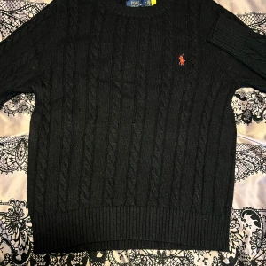 Svart stickad tröja från Ralph Lauren - Säljer en klassisk svart stickad tröja från Ralph Lauren med kabelstickat mönster. Tröjan har en liten röd logga på bröstet och ribbade muddar vid ärmslut och nederkant. Perfekt för kyligare dagar!