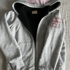 Grå hoodie från Zadig & Voltaire - Säljer en grå hoodie från Zadig & Voltaire med dragkedja och fodrad med mjuk fleece. Den har en stor luva med röd text och en tryckt etikett på ena ärmen. Storlek 150/S