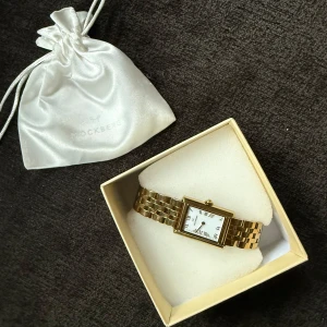 Guldfärgad klocka från Mockberg - Elegant guldfärgad klocka från Mockberg med rektangulär urtavla och romerska siffror. Armbandet är i metall och kommer med extra länkar för justering. Perfekt för en stilren look.