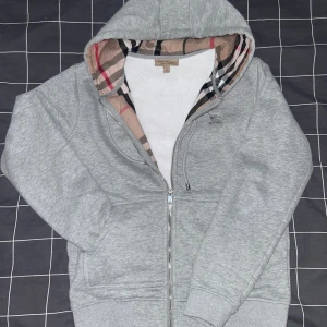 Grå huvtröja med mönstrat foder - Säljer en grå huvtröja från Burberry med dragkedja och ett ikoniskt rutigt foder i huvan. Tröjan har en broderad logga på bröstet och är tillverkad i mjukt bomullsmaterial. Perfekt för en stilren och bekväm look.